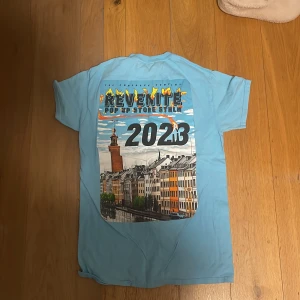 Blå Revenite  Pop Up t-shirt S - Ljusblå t-shirt från Revenite  Pop Up Store Sthlm 2023 med stor färgglad print på ryggen som visar Stockholms stadshus och husfasader. Klassisk rund hals och korta ärmar. Skön och mjuk i bomull, perfekt för dig som gillar streetwear och unika tryck.
