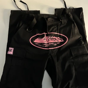 Svarta cargopants Corteiz XS - Svarta cargopants från Corteiz med rosa tryck och patchdetaljer. Byxorna har stora fickor på benen och justerbara band. Materialet är 65% bomull och 35% polyester. 
