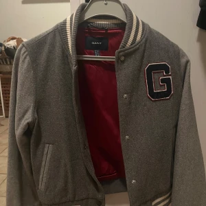 Grå bomberjacka från Gant med G-logga - Snygg grå bomberjacka från Gant med broderad G-logga på bröstet. Jackan har ribbade muddar och krage med vita ränder, röd insida och knäppning med tryckknappar. Perfekt för dig som gillar klassisk college-stil. Nypris 3000