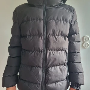 Svart pufferjacka från Superdry - Säljer en svart pufferjacka från Superdry. Endast använd fåtal gånger förra vintern, nyskick! Avslappnad passform, inte för smal, inte för vid utan precis lagom.  Hållbar vattenavstötningsförmåga, det här materialet har belagts med en avancerad vattentålig yta Resårsnodd i huvan Dragkedja fram Dubbla fickor med dragkedjor Ärmslut med resår Resårsnodd i fållen Fleecefodrad överkroppsdel Invändig meshficka Broderad Superdry-logga på baksidan och ärmen.