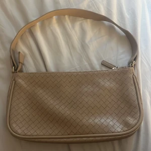 Beige axelväska i flätad skinnimitation - Snygg beige axelväska med flätad design och dragkedja upptill. Väskan har både en bred axelrem och en justerbar, avtagbar lång rem. Tillverkad i skinnimitation och rymmer det viktigaste för dagen. Perfekt för dig som gillar stilrena detaljer.