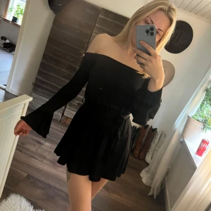 Svart offshoulder jumpsuit - Supersnygg svart byxdress med offshoulder och långa utsvängda ärmar. Klänningen har ett knytband i midjan som framhäver figuren och ger en trendig look. Perfekt för dig som gillar stilrena och coola outfits. Stretchiga material så passar både xs och s