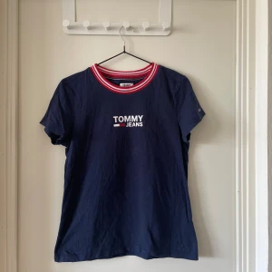 Blå Tommy Jeans t-shirt med röd/vit krage - Mörkblå t-shirt från Tommy Jeans i bomull med korta ärmar och ribbad rund halsringning i rött och vitt. Framsidan har klassisk Tommy Jeans-logga i vitt, rött och blått. Perfekt för en sportig och avslappnad look.