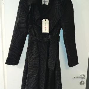 Svart zebramönstrad kappa från H&M - Svart kappa från H&M med zebramönster och bälte i midjan. Kappan har långa ärmar och en snygg omlottkrage. Materialet är mjukt och har en lätt glansig finish som ger en cool look.