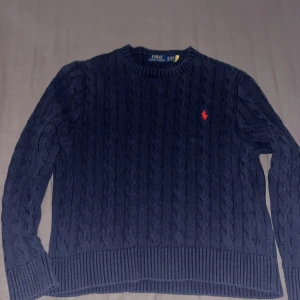 Mörkblå kabelstickad tröja  - Hej säljer min mörkblå polo tröja med klassisk rund hals och röd broderad logga på bröstet. Tröjan har långa ärmar och ribbade muddar vid ärmslut och nederkant. Perfekt nu inför vintern.