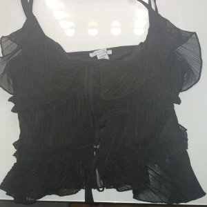 Svart volangig linnetopp från AMISU - Supersöt svart linnetopp från AMISU med tunna axelband, volanger och plisserad mesh. Toppen har knytning framtill och är lätt transparent med romantiska detaljer. Perfekt för dig som gillar en feminin och trendig look.