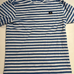 Randig t-shirt från J.Lindeberg - Fräsch t-shirt från J.Lindeberg i blått och vitt med klassiskt randigt mönster. T-shirten har rund hals, korta ärmar och en liten svart logotyp på bröstet. Tillverkad i mjuk bomull som känns skön mot huden. Perfekt för dig som gillar stilren och enkel design.