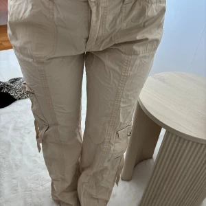 Beiga cargopants från Gina Tricot - Säljer ett par beiga cargopants från Gina Tricot i storlek 36. Byxorna har flera fickor med lock och coola detaljer som snörning på benen. De är tillverkade i ett lätt och syntetiskt material och har en avslappnad passform med resår i midjan.