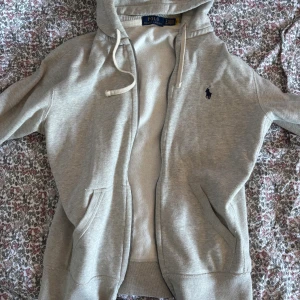 Grå hoodie från Polo Ralph Lauren - Grå hoodie från Polo Ralph Lauren med dragkedja, huva och snörning. Klassisk broderad logga på bröstet och två fickor framtill. Mjuk och skön bomullsblandning, perfekt för chill dagar.