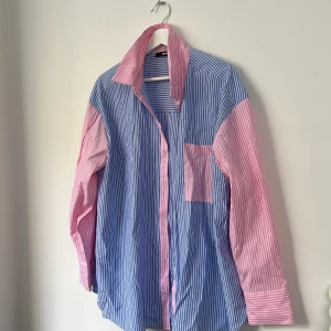 Randig skjorta från BikBok i blått och rosa - Säljer en oversized skjorta från BikBok med blå och rosa ränder. Skjortan har lång ärm, klassisk krage och bröstficka. Tillverkad i en mix av polyester och bomull. Perfekt för en chill och färgglad look.