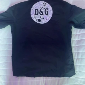 Svart skjorta från Dolce & Gabbana - Svart skjorta från D&G med stort tryck på ryggen och klassisk krage. Skjortan har knappar framtill och korta ärmar. Snygg och stilren med tydlig logga på ryggen, perfekt för dig som gillar exklusiva märken.