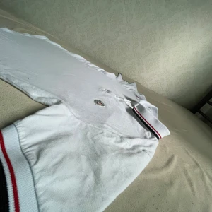 Vit pikétröja från Moncler S - Vit pikétröja från Moncler med klassisk krage och knäppning framtill. Tröjan har Moncler-logga broderad på bröstet och detaljer i rött och marinblått på krage och ärmslut. Tillverkad i bomull och har korta ärmar. Perfekt för en clean och sportig look.