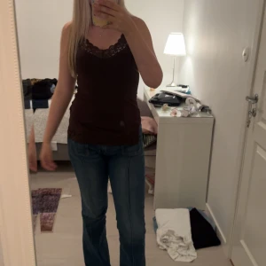 Brun linnetopp med spetsdetalj - Snygg brun linnetopp med smala axelband och v-ringning dekorerad med spets. Toppen har en figurnära passform och är tillverkad i mjukt bomullsmaterial. Perfekt för dig som gillar en enkel men ändå söt stil.