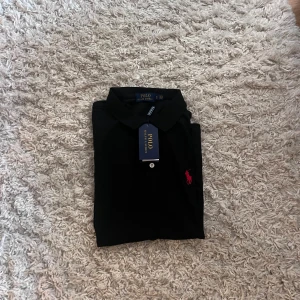 Svart pikétröja från Ralph Lauren - Svart pikétröja från Polo Ralph Lauren med klassisk krage, korta ärmar och knappar framtill. Ikoniska röda broderade loggan på bröstet. Tillverkad i mjuk bomull för en skön känsla. Perfekt för en clean och stilren look.