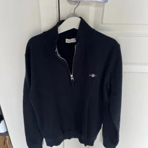 Svart stickad tröja från Gant - Svart stickad tröja från Gant med halv dragkedja och broderad logga på bröstet. Tröjan har hög krage och långa ärmar, perfekt för kyliga dagar. Klassisk och stilren design som funkar till det mesta.