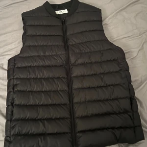 Svart dunväst från H&M - Svart dunväst från H&M med quiltad design och dragkedja framtill. Västen har en klassisk, rak passform och är perfekt för lager-på-lager-stil. Enkel och stilren look som funkar till många outfits.