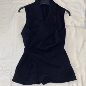 Svart omlottväst med krage - Stilren svart väst med omlottknäppning och bred krage. Västen har en lätt strukturerad yta och knytband i sidan för en snygg passform. Perfekt att styla över en skjorta eller t-shirt för en cool look.