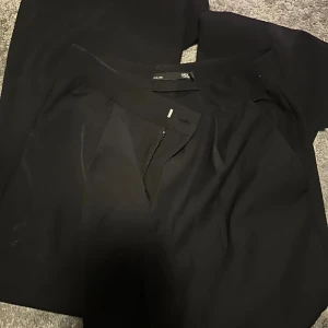 Svarta kostymbyxor från 157 - Svarta kostymbyxor från 157 med klassisk passform och dragkedja framtill. Byxorna har en stilren look och är tillverkade i ett mjukt tyg som känns bekvämt. Perfekta för dig som gillar en clean och enkel stil.