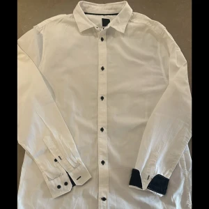 Vit slim fit skjorta från H&M - Vit skjorta från H&M i slim fit-modell med svarta knappar och svarta detaljer på manschetterna. Klassisk krage och långärmad design, perfekt för en clean och stilren look. Skjortan är tillverkad i bomull och passar dig som gillar en smalare passform.