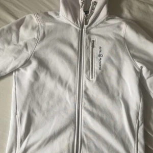 Vit fleecejacka från Sail Racing - Säljer en vit fleecejacka från Sail Racing med huva och hel dragkedja. Jackan har en bröstficka med dragkedja och logga på bröstet. Mjuk insida och stilren design, perfekt för lager-på-lager. Passar dig som gillar sportig och clean stil. S/M storlek