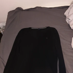 Svart v-ringad tröja Tommy Hilfiger - Svart långärmad tröja från Tommy Hilfiger med v-ringning och liten broderad logga på bröstet. Klassisk och stilren modell i mjukt bomullsmaterial, perfekt för dig som gillar enkel och clean stil.