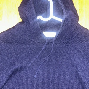 Mörkblå stickad hoodie med huva - Mörkblå stickad hoodie med huva och snörning. Tröjan har lång ärm och en klassisk passform, perfekt för kyliga dagar. Enkel och stilren design som funkar till det mesta.