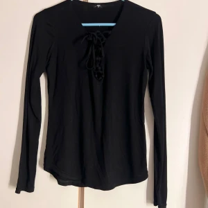Svart långärmad topp med snörning - Svart långärmad topp med v-ringning och snygg snörningsdetalj framtill. Modellen är figurnära och enkel, perfekt att styla med jeans eller kjol. Mjukt och stretchigt material som känns skönt mot huden.