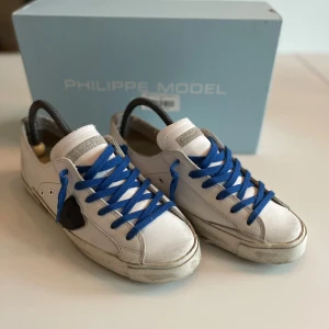 Philippe Model - Snygga vita Philippe Model sneakers med blå snören och svarta detaljer. Skorna har en klassisk låg profil, rund tå och platt sula med tydlig logga undertill. Tillverkade i skinn med mockadetaljer och cool patch på sidan. Skick 10/10 bara använt dom 15 gånger! 