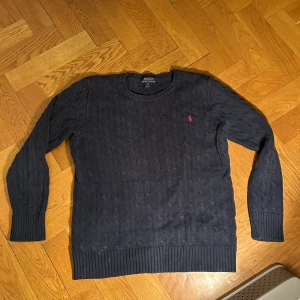 Mörkblå stickad tröja från Polo Ralph Lauren - Snygg mörkblå stickad tröja från Polo Ralph Lauren med klassiskt kabelmönster och röd broderad logga på bröstet. Rund halsringning och ribbade muddar vid ärmslut och nederkant. 