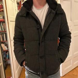 Mörkgrå pufferjacka med huva - Snygg mörkgrå pufferjacka från Pier One. Jackan har både dragkedja och knappar framtill, samt inner ficka. Jackan har en modern, normal passform. Perfekt för kyliga dagar.