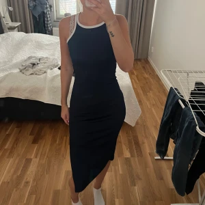 Marinblå ribbad klänning - Snygg marinblå klänning med vit kant runt halsen och ärmhålen. Klänningen är ribbad och har en tight passform med slits på sidan. Axelbandslös design med spetsdetaljer på axelbanden ger en fräsch och modern look. Perfekt för dig som gillar stilrena och sportiga vibes.