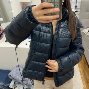 Mörkblå pufferjacka med huva - Säljer en mörkblå pufferjacka med avtagbar huva och dragkedja framtill. Jackan har en quiltad look, två fickor med dragkedja och är långärmad. Perfekt för kalla dagar och riktigt skön att bära.knappt använd 