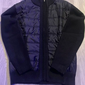 Svart cardigan jacka med stickade ärmar - Svart jacka med quiltad framsida i blankt material och stickade ärmar. Insidan har ett grått camouflagefoder i fleece som gör den extra mysig. Jackan har dragkedja framtill och ribbade muddar. Perfekt för dig som vill ha en stilren och bekväm look.