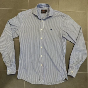 Randig skjorta från Morris  - Snygg blåvit randig skjorta från Morris Stockholm med klassisk krage och broderad logga på bröstet. Skjortan har långa ärmar, vita knappar och är tillverkad i mjuk bomull. Perfekt för dig som gillar stilrena och tidlösa plagg.