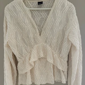 Cremevit volangblus från Gina Tricot - Supersöt vit blus från Gina Tricot i transparent, vågigt strukturerat tyg. Blusen har v-ringning, långa ärmar och volangdetaljer framtill och baktill. Även lite i armarna (längst ner) Perfekt att styla med jeans eller kjol för en trendig look.