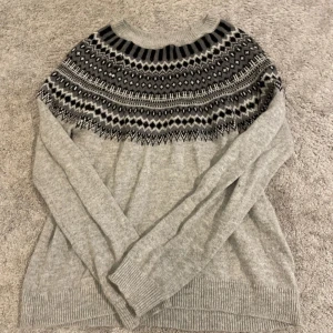 Grå stickad tröja med mönster L.O.G.G. - Säljer en grå stickad tröja från L.O.G.G. med snyggt svartvitt mönster runt halsen. Tröjan har rund halsringning, långa ärmar och ribbade muddar. Perfekt för kyliga dagar och ger en mysig vibe.