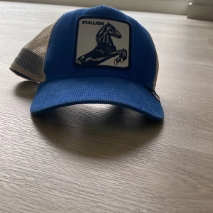 Blå keps Goorin Bros Stallion - Snygg blå keps från Goorin Bros med vit mesh baktill och patch med svart hästmotiv och texten 'Stallion' framtill. Kepsen är från The Farm-serien och har justerbar snapback. Materialet är 57% polyester och 43% bomull.