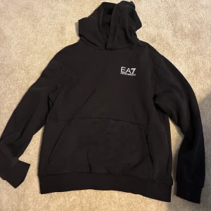 Svart hoodie från Emporio Armani EA7 - Svart hoodie från Emporio Armani EA7 med vit logga på bröstet. Klassisk känguruficka framtill och huva. Perfekt för dig som gillar stilrena och exklusiva streetwear-plagg.