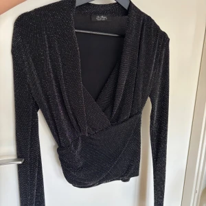 Glittrig omlottopp från New Mission - Svart omlottopp från New Mission med glittriga silverdetaljer. Toppen har lång ärm, djup v-ringning och en snygg drapering i midjan. Perfekt för dig som vill sticka ut med en festlig look. Materialet är stretchigt och följsamt.