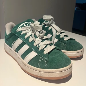 Adidas Campus gröna sneakers mocka - Säljer ett par gröna Adidas Campus sneakers i mocka med klassiska vita ränder och vit sula. Skorna har rund tå, platt sula och snörning. Adidas logga syns på både häl och plös. Perfekta för dig som gillar retrostil och streetwear.