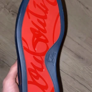 Mörkblå mockasneakers från Louboutin - Säljer ett par exklusiva sneakers från Christian Louboutin i mörkblå mocka med ikonisk röd sula och snörning. Skorna har en stilren design med rund tå och diskreta sömmar, perfekta för dig som vill sticka ut med lyxiga detaljer.