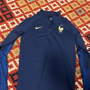 Blå långärmad Nike FFF Dri-Fit tröja - Säljer en marinblå långärmad fotbollströja från Nike med FFF-logga och två stjärnor på bröstet. Slim fit-modell i Dri-Fit material som andas, med halv dragkedja framtill och guldigt Nike-märke. Snygga blå detaljer på ärmarna.