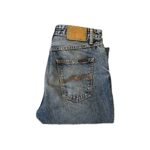 Blå jeans från Nudie Jeans - Snygga blå jeans från Nudie Jeans med klassisk femficksdesign och orangea sömmar på bakfickan. Jeansen har rak passform och normal midja.