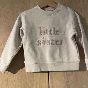 Beige sweatshirt från LIVLY, 92 - Supersöt beige sweatshirt från LIVLY med broderad text 'little sister' i ljusrosa framtill. Tröjan har rund hals, långa ärmar och praktiska knappar på axeln. Perfekt för småsyskon som vill vara extra gulliga.