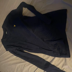 Mörkblå långärmad tröja Lyle & Scott - Mörkblå långärmad tröja från Lyle & Scott Junior med klassisk gul logga broderad på bröstet. Tröjan har rund halsringning och är tillverkad i mjuk bomull, perfekt för en clean och stilren look.