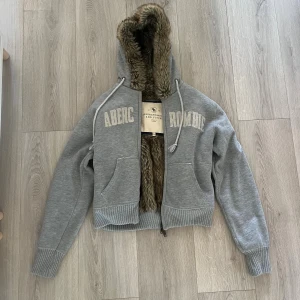 Äkta vintage Abercrombie & Fitch fur zip hoodie - Grå hoodie från Abercrombie & Fitch med dragkedja, stor huva och mysigt fuskpälsfoder. Broderad logga framtill, ribbade muddar och snörning i huvan. Perfekt för kalla dagar när du vill vara både varm och snygg.       Tar gärna fler bilder om det behövs, svarar snabbt på frågor! Den är äkta så om du vill ha bild på ngt speciellt för att bekräfta det, går det att ordna!🩶🍂