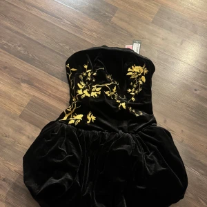 Svart sammetsklänning PrettyLittleThing - Supersnygg svart sammetsklänning från PrettyLittleThing med guldig broderad blomdetalj upptill. Axelbandslös modell med puffig kjol och figurnära passform upptill. Perfekt för dig som vill sticka ut med lyxiga detaljer och cool siluett.