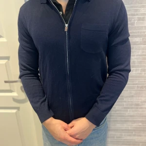 Mörkblå merinoullskofta från Lindbergh - Snygg mörkblå kofta från Lindbergh i mjuk merinoull. Modellen har dragkedja framtill, bröstficka och klassisk krage. Perfekt för lager-på-lager och passar till jeans eller chinos. Långärmad och med en clean, stilren look. Storlek m, hör av dig vid frågor