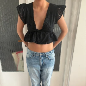 Svart blus med broderade hål och volang - Säljer en svart blus med djup v-ringning och broderade hål i blommönster. Toppen är croppad med volang nertill och har korta, vingeformade ärmar. Perfekt för dig som gillar en luftig och trendig look. Har ingen lapp vet inte vart den är ifrån. Skulle säga xs eller S. 