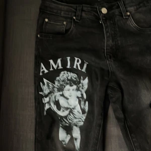 Svarta Amiri jeans med ängeltryck - Säljer ett par svarta jeans från Amiri med stort vitt ängeltryck och logga på ena benet. Klassisk femficksmodell med snygga detaljer och metallmärke bak. Jeansen har en modern passform och är tillverkade i jeansmaterial.